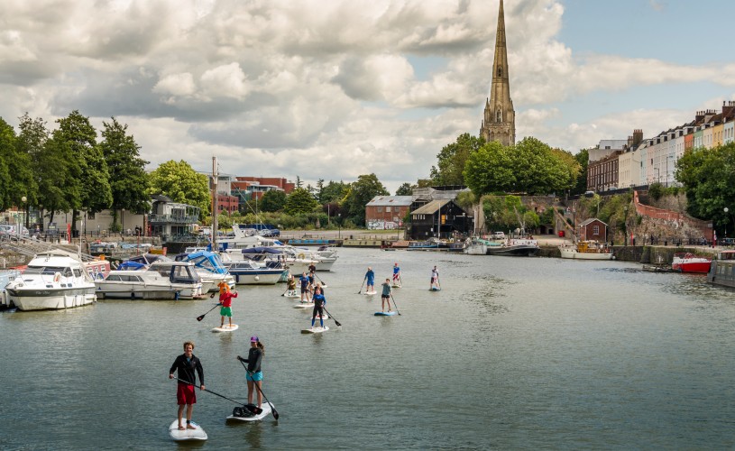 People stand up paddleboarding with boats and St Mary Redcliffe in the background